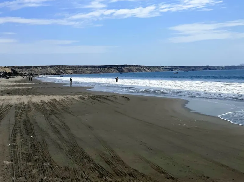 Playa ancha de arena oscura con olas suaves y gente caminando a lo lejos