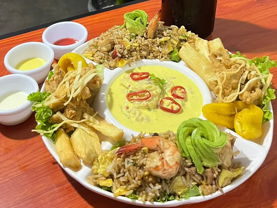 Fuente blanca con arroz chaufa de langostino, pescado frito con yuca y plátano, y una crema amarilla con ají rojo al centro